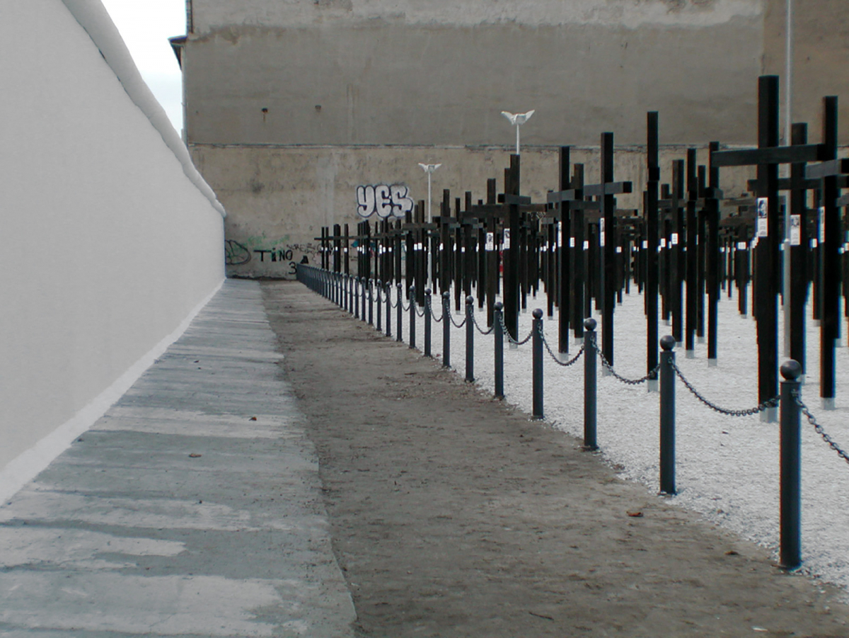 Schwarze Kreuze auf einer der beiden Brachflächen, ihnen gegenüber die wiederaufgebauten weißen Segmente der Mauer.