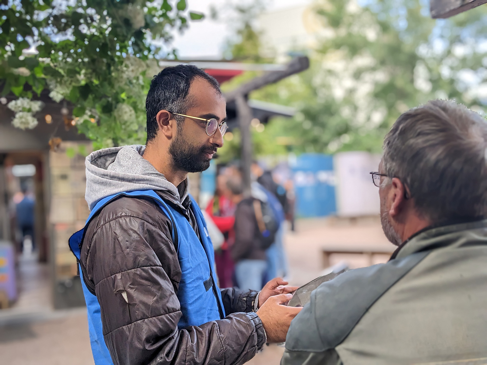 Ein junger Mann in blauer Weste und mit einem Tablet in der Hand befragt einen Touristen.ge" und einer stilisierten weißen Zeichnung des Ortes.