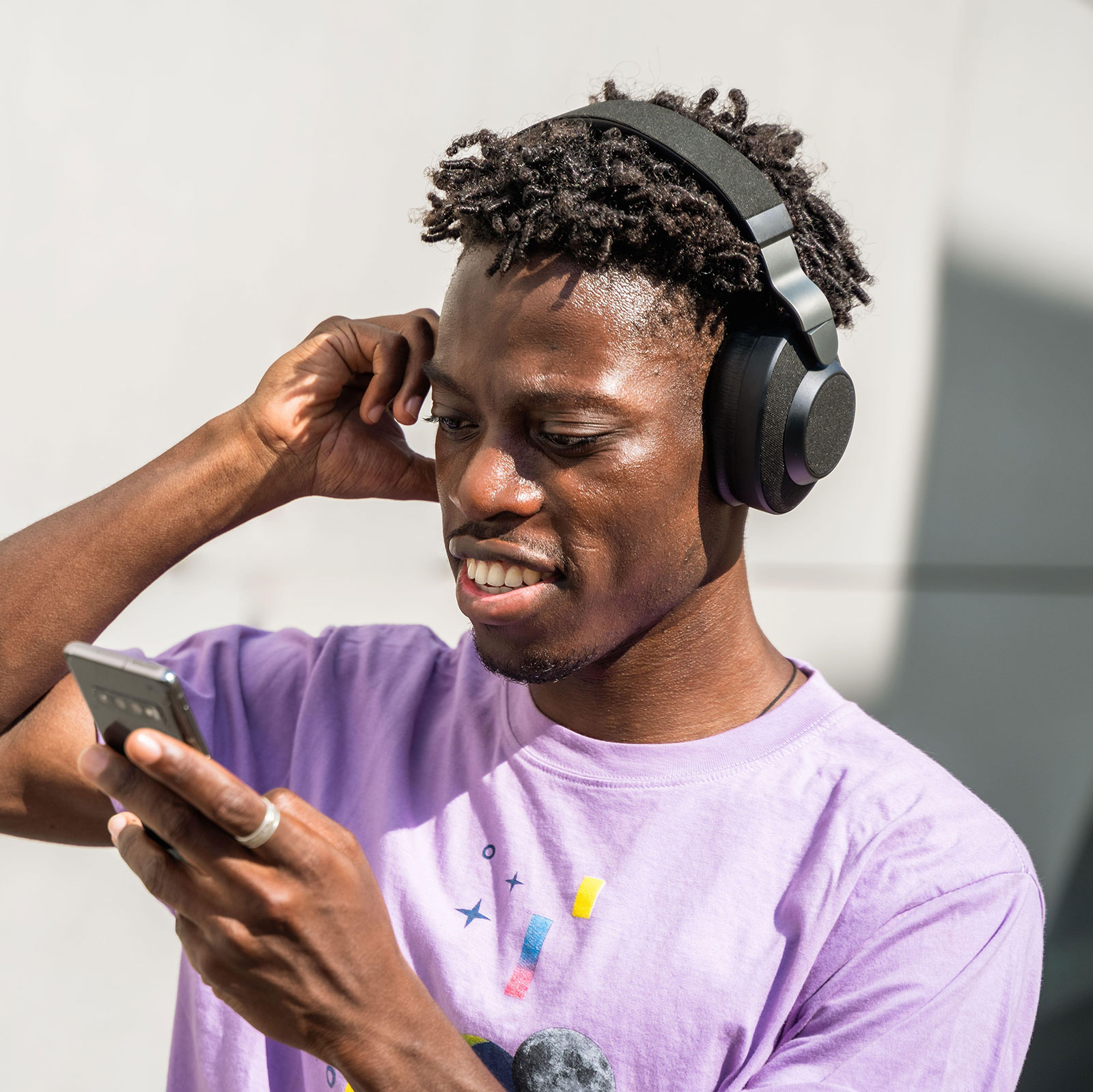 Ein junger schwarzer Mann in lila Shirt mit kurzen Dreadlocks schaut interessiert auf sein Handy und trägt over-ear Kopfhörer.