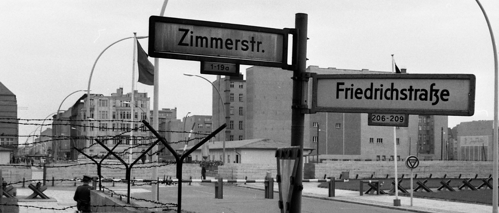 Straßenschild Zimmer- Ecke Friedrichstraße, dahinter eine Steinmauer mit Stacheldraht und Schlagbäume des DDR-Grenzübergangs.